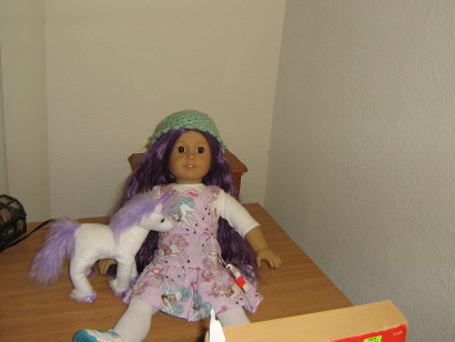 On a small dresser sits an 18" AG doll with LONG PURPLE HAIR is on the dresser! She wears a unicorn-print bodice and skirt, white socks, tennis shoes, a shirt underneath, and a turquoise crocheted hat. A white unicorn with purple hair stands beside her.